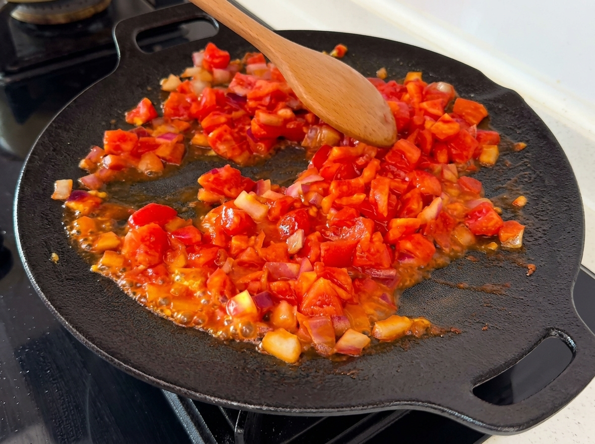 Smashed and diced tomatoes mixing with onions on a hot pan, showing visible juices bubbling.