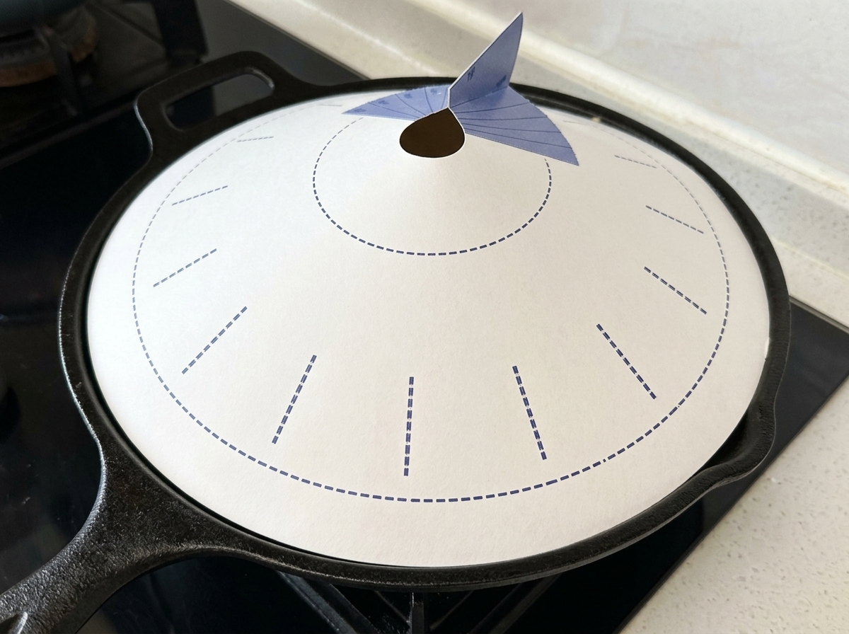A black cast iron pan covered with a white conical paper lid featuring blue markings, sitting on a stovetop.