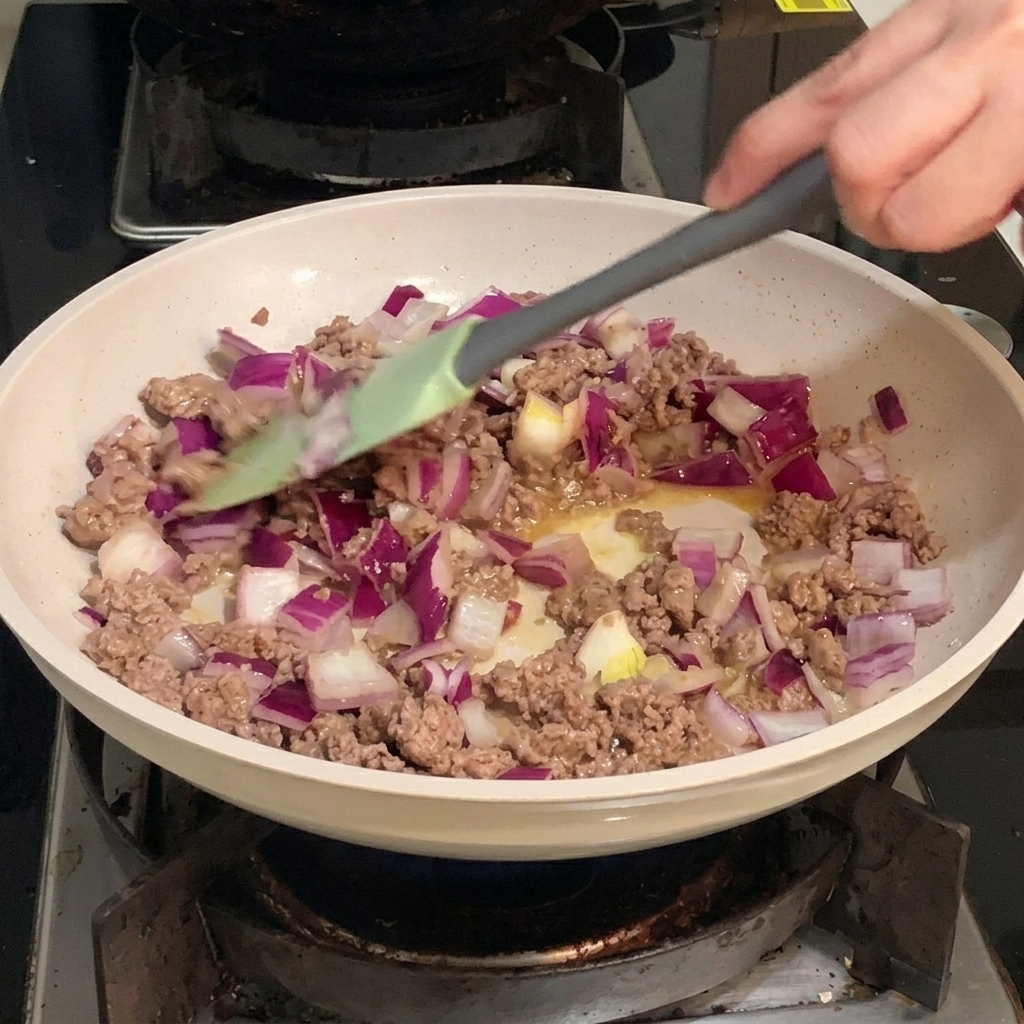Diced pieces of purple and white red onion being tossed with browned ground beef in a frying pan.
