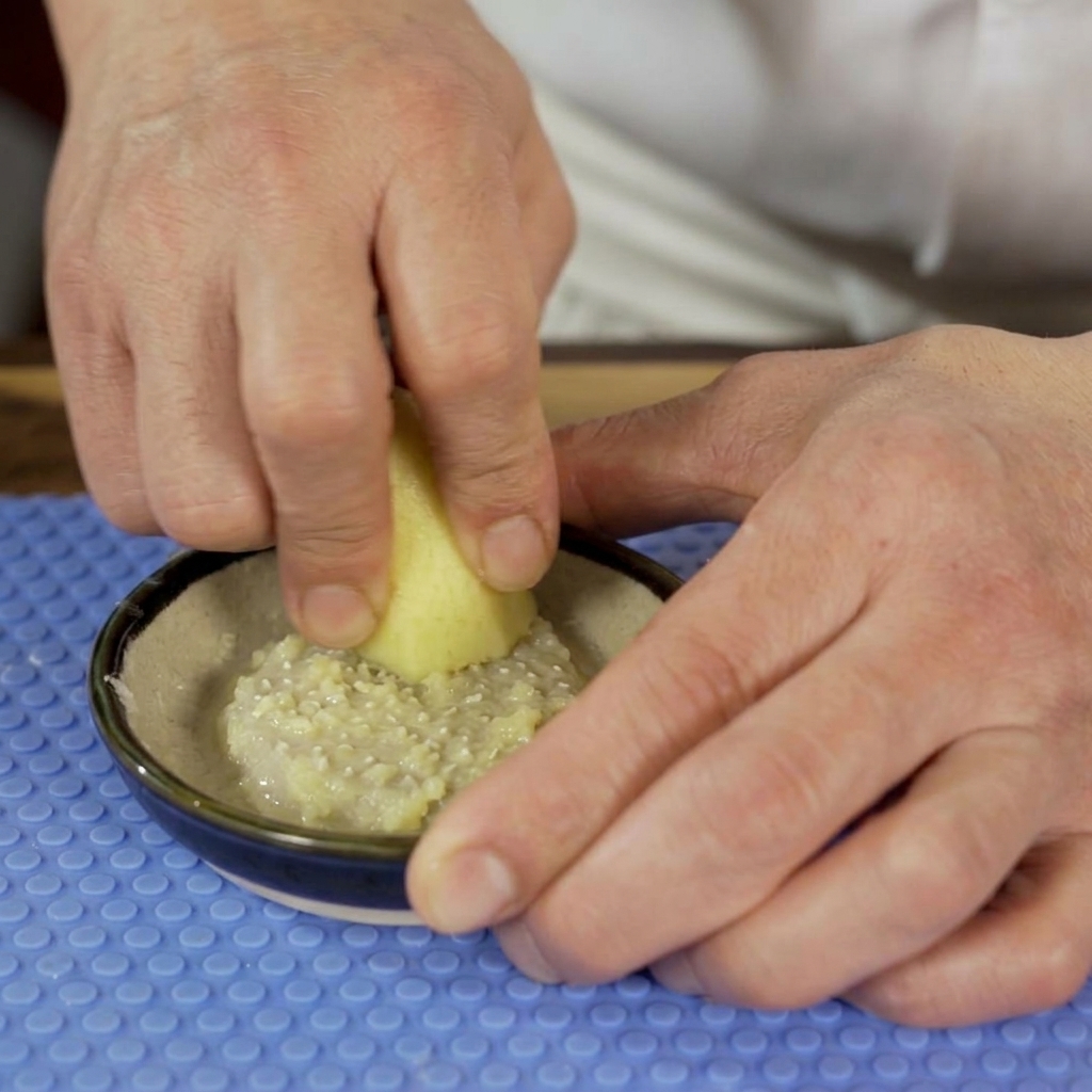 A person using a ceramic grater to turn a piece of fresh ginger root into a smooth, wet paste in a small bowl.