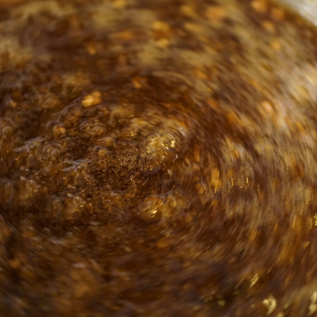 A close-up shot of a dark brown sauce bubbling vigorously in a pot while being stirred rapidly.
