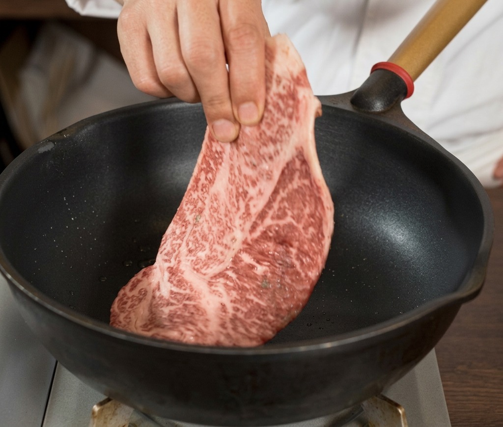 A hand placing a large, well-marbled raw steak seasoned with black pepper into a hot black skillet.