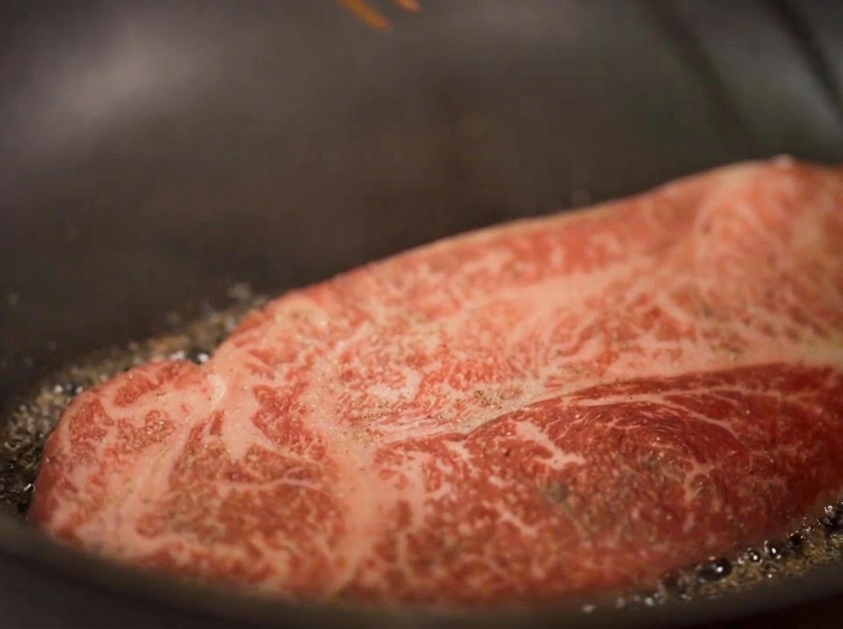 A close-up shot of a thick steak searing in oil, with visible steam rising and the bottom edges turning golden brown.