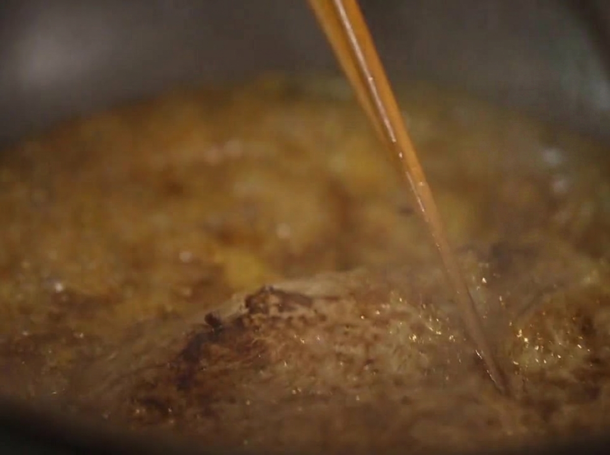 A close-up of a steak simmering in a thick, bubbling brown sauce, being basted with a pair of chopsticks.