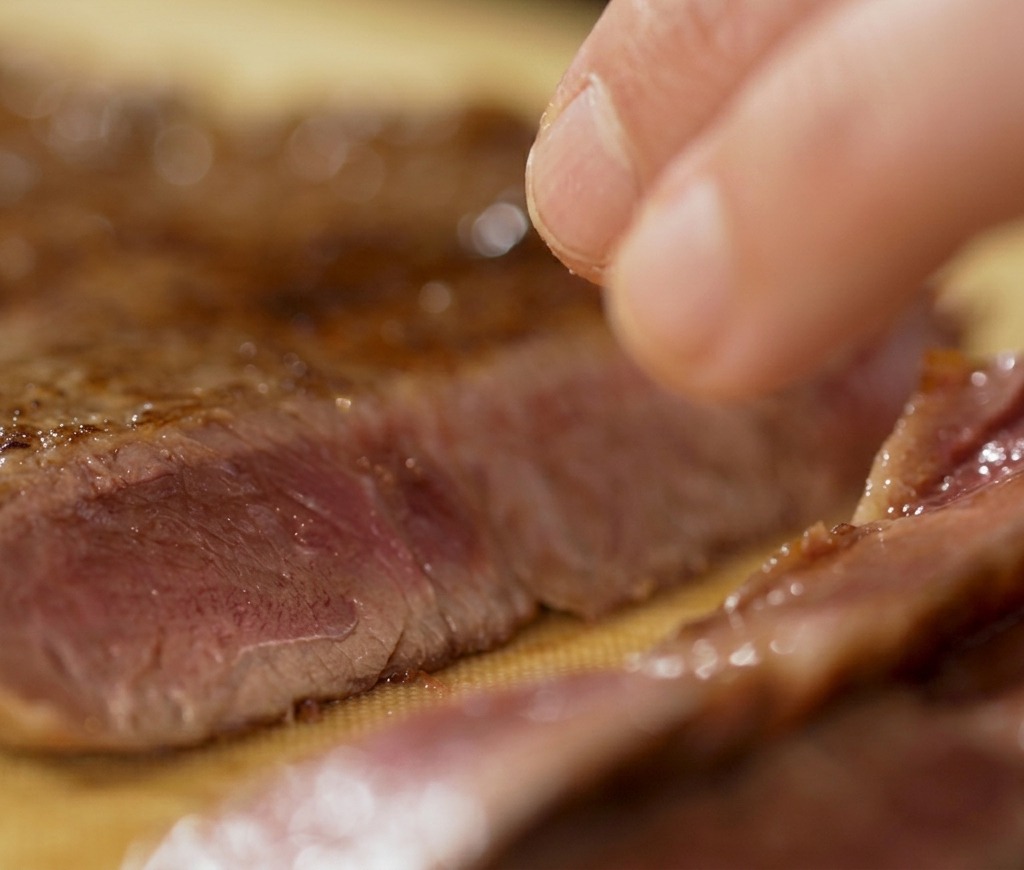 A hand using a knife to slice a piece of seared beef on a cutting board, showing a perfectly cooked pink interior.