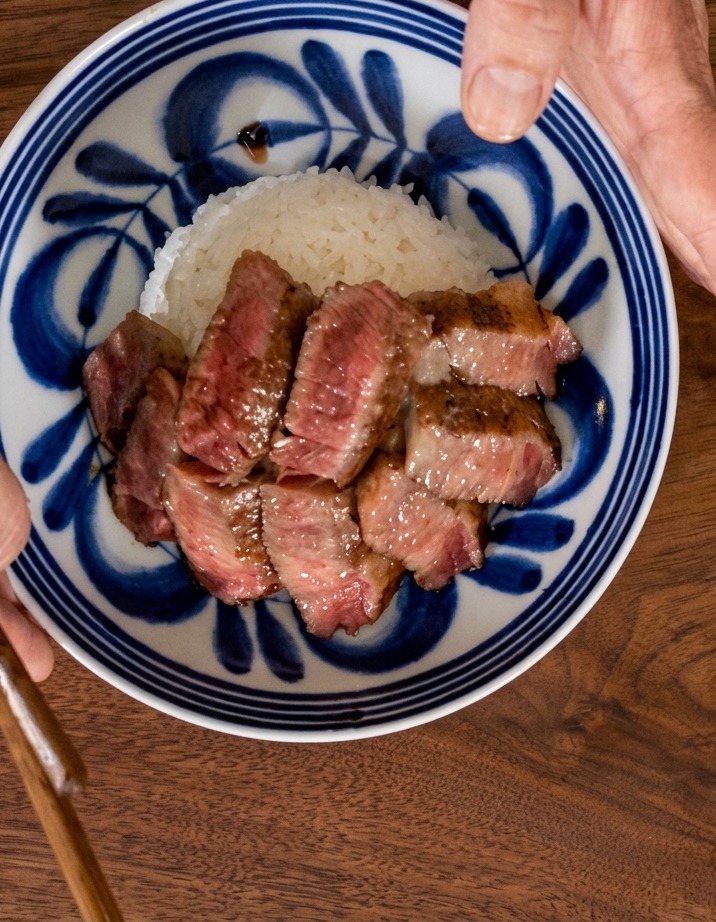 A top-down view of a hand holding a blue patterned bowl as steak slices are fanned out across a bed of white rice.