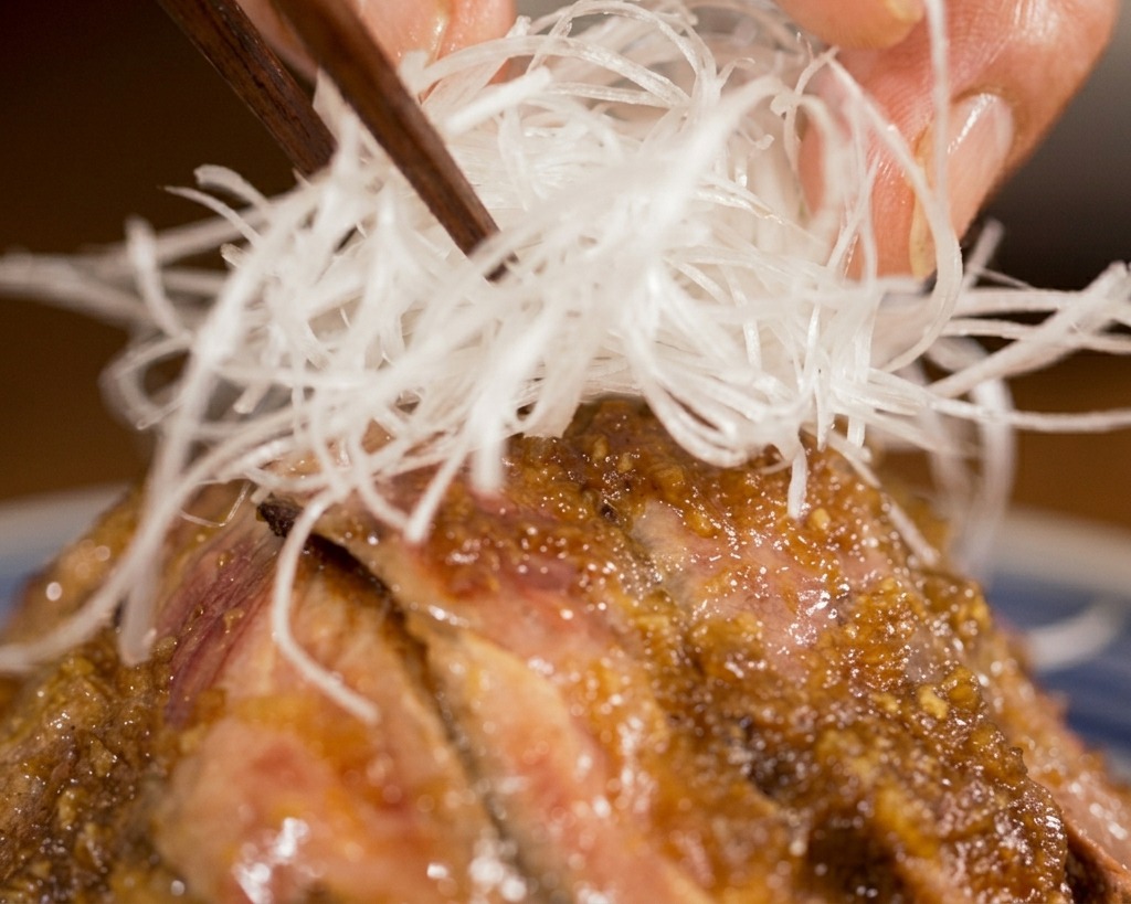 Chopsticks placing a heap of thin white leek shreds onto the center of a sauced steak donburi bowl.