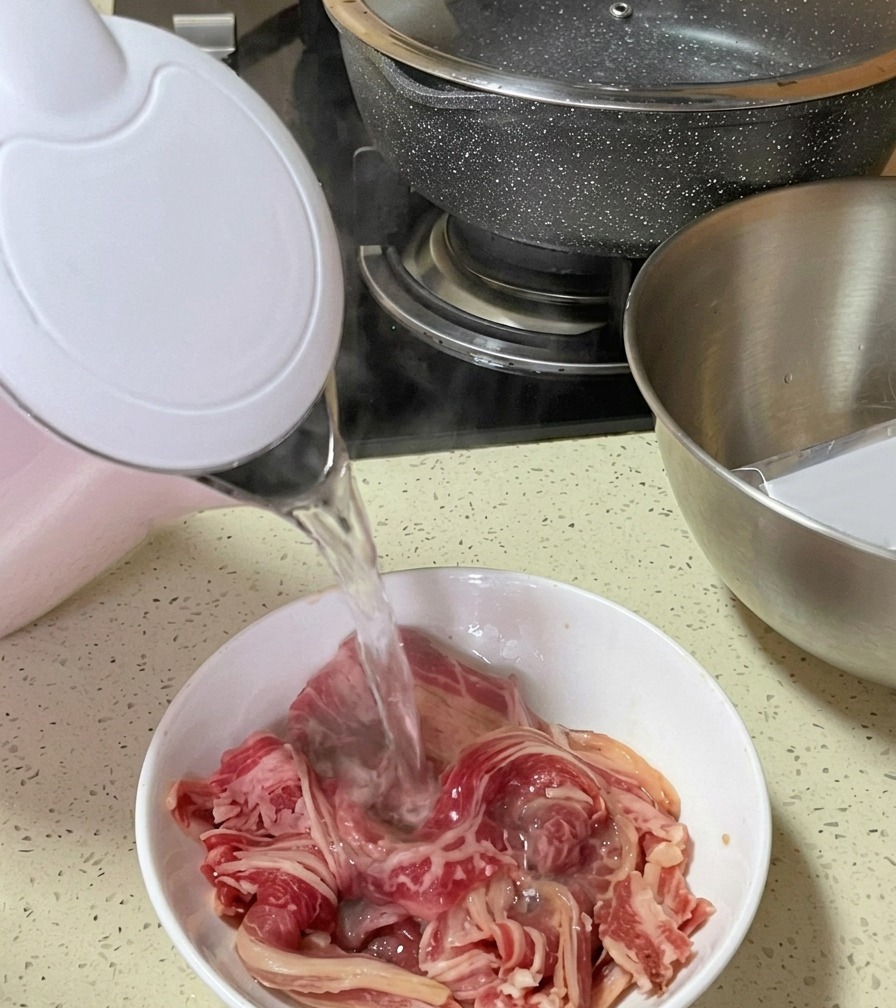 Steaming boiling water being poured from a pink and white electric kettle onto raw, thinly sliced beef strips in a bowl.