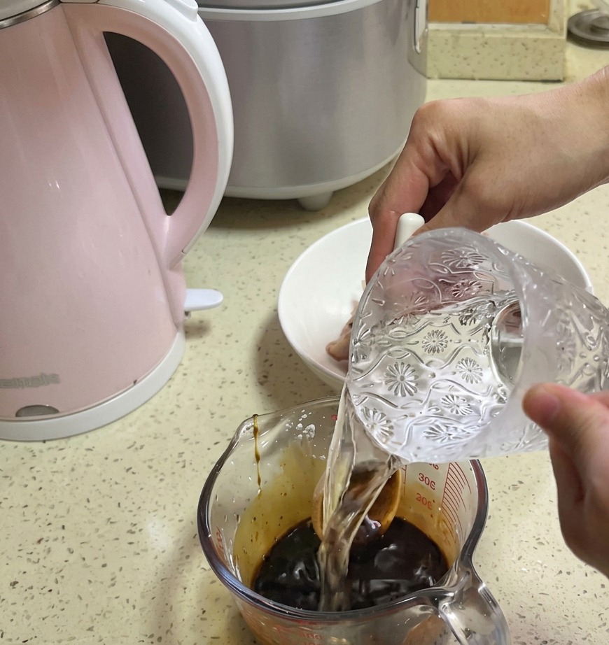 Clear water being poured from a decorative glass pitcher into a measuring cup containing a dark brown sauce base.