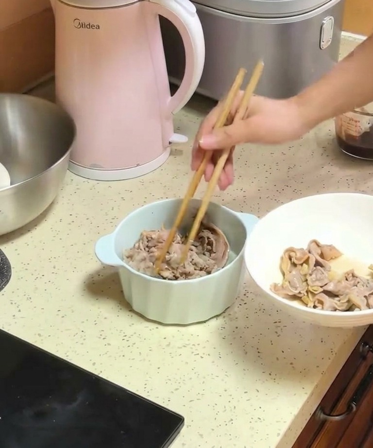 A person using wooden chopsticks to move blanched beef strips from a white plate into a bowl containing cooked onions.