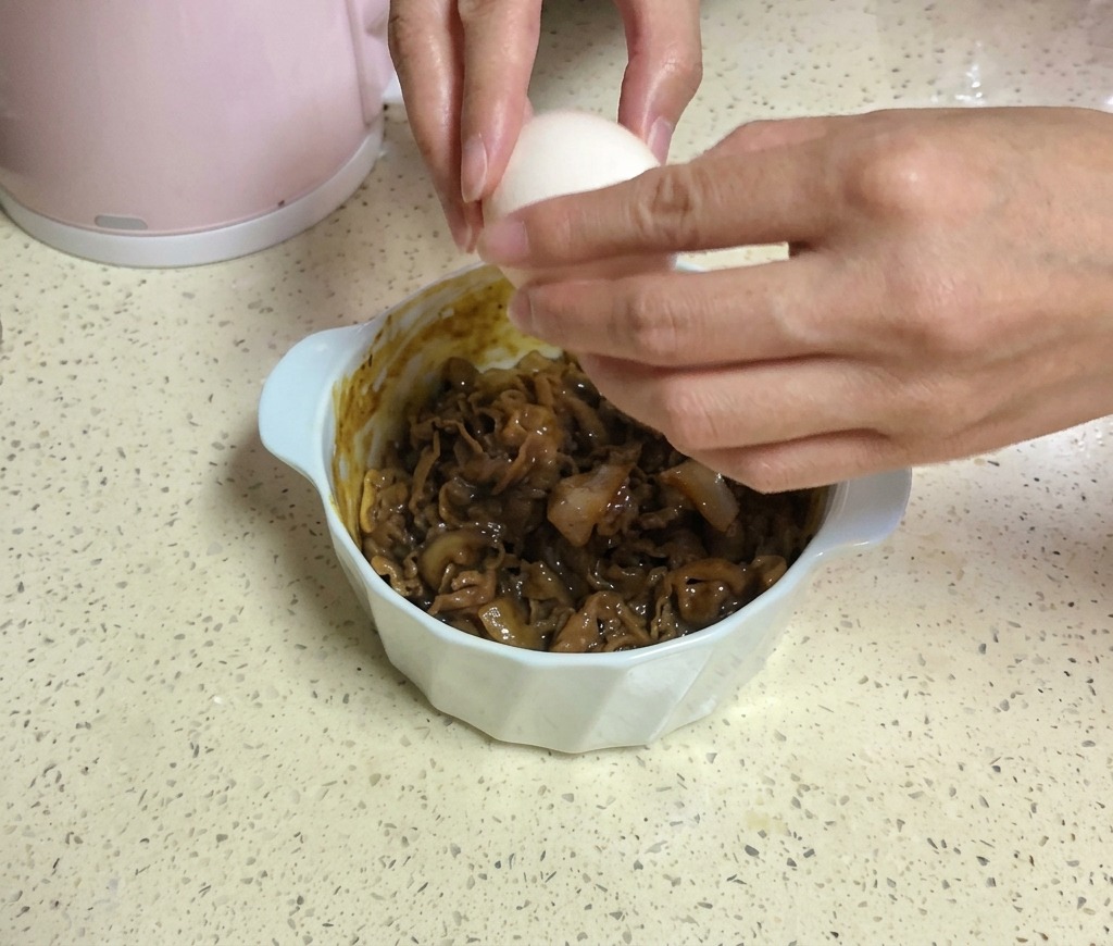 Two hands cracking a white egg over a bowl of beef topped with two slices of yellow cheese.