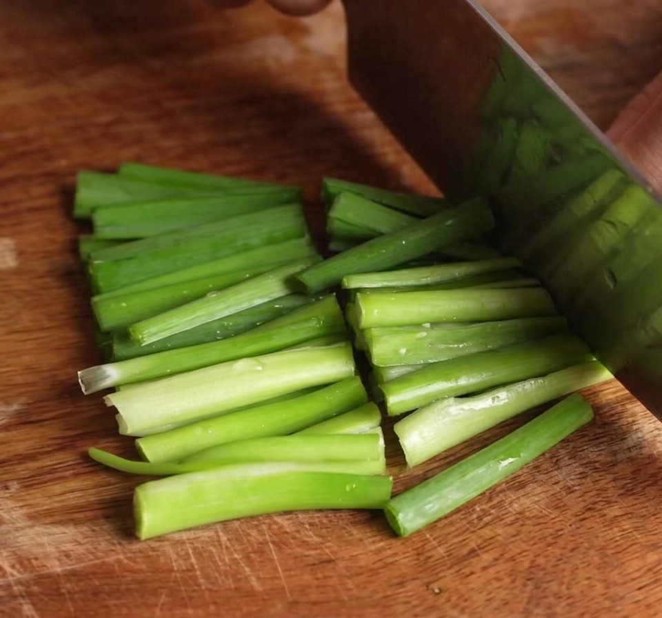 A knife slicing through a bundle of bright green chives on a wooden cutting board, creating short segments.