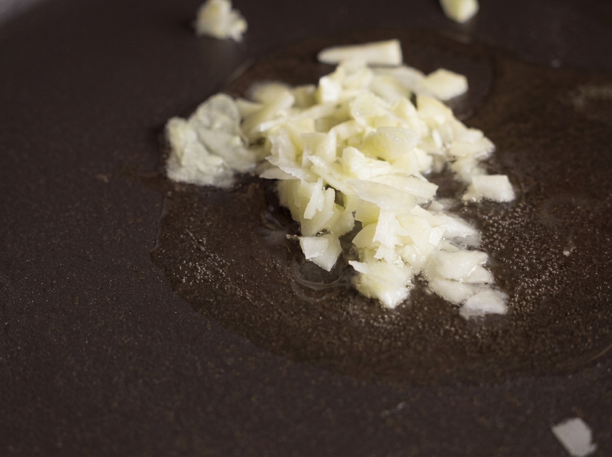 Finely minced garlic sizzling in a pool of clear oil in a dark non-stick frying pan.