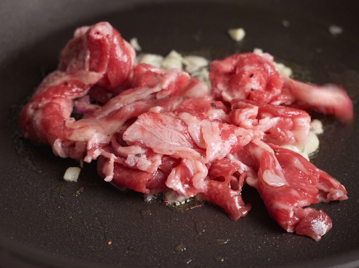Slices of raw beef being added to a pan containing sautéed garlic, with some parts of the meat beginning to turn brown from the heat.