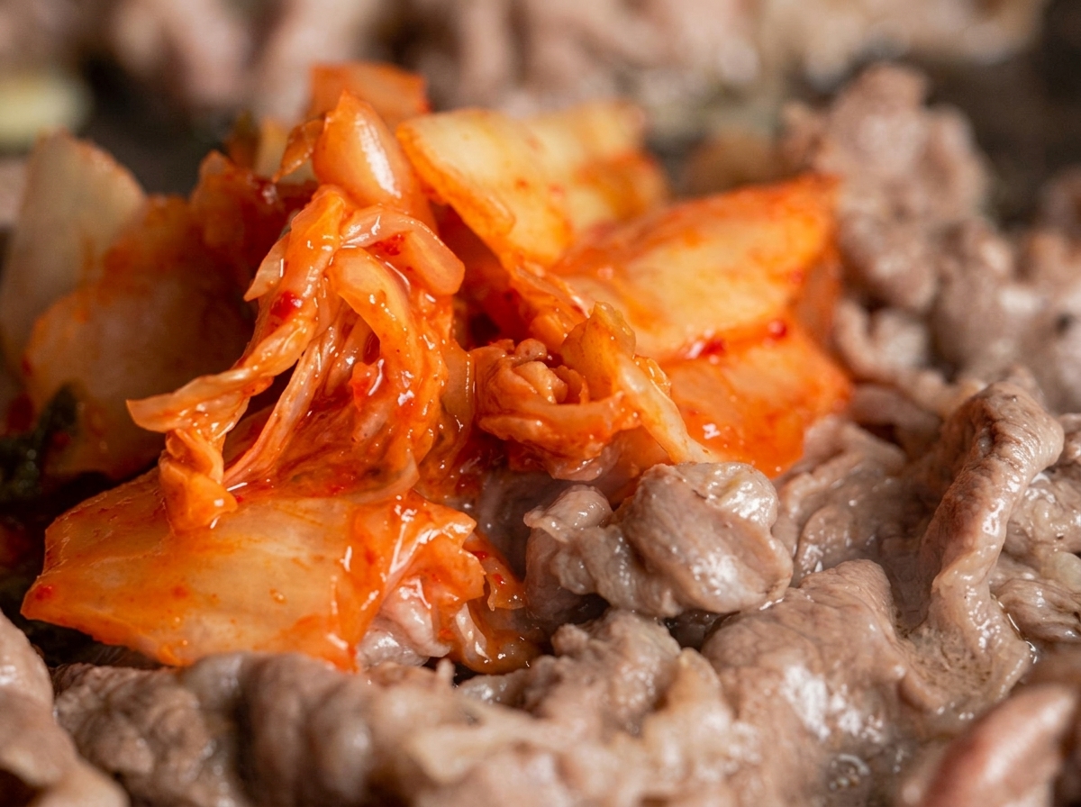 A heap of vibrant red kimchi being mixed into thinly sliced beef strips in a frying pan.