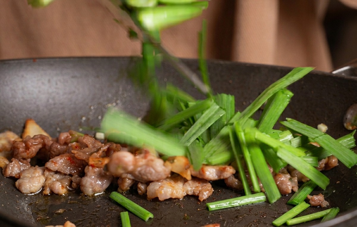 A hand dropping chopped green chives into a pan containing a stir-fry of beef and kimchi.
