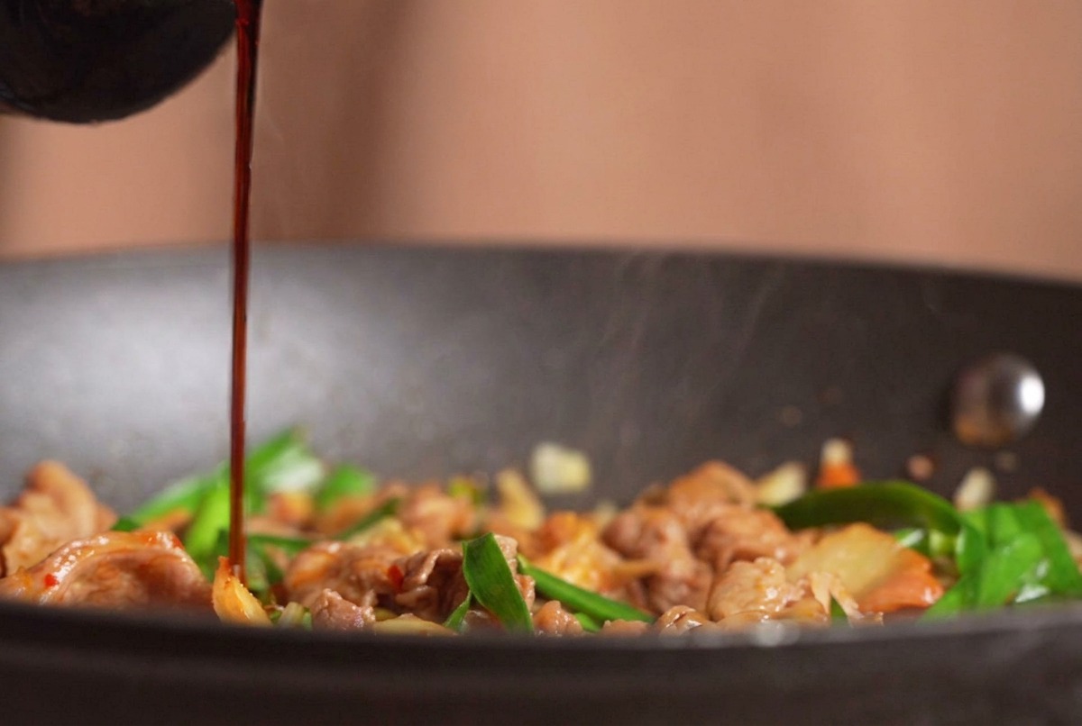 Dark soy sauce being poured from a bottle onto a steaming stir-fry of beef, kimchi, and chives.
