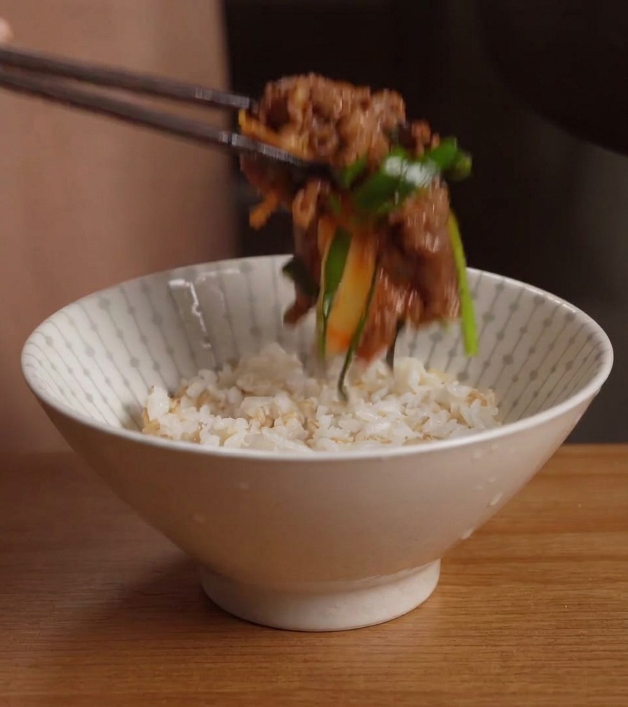 Chopsticks placing a serving of stir-fried beef and kimchi onto a white bowl filled with cooked rice.