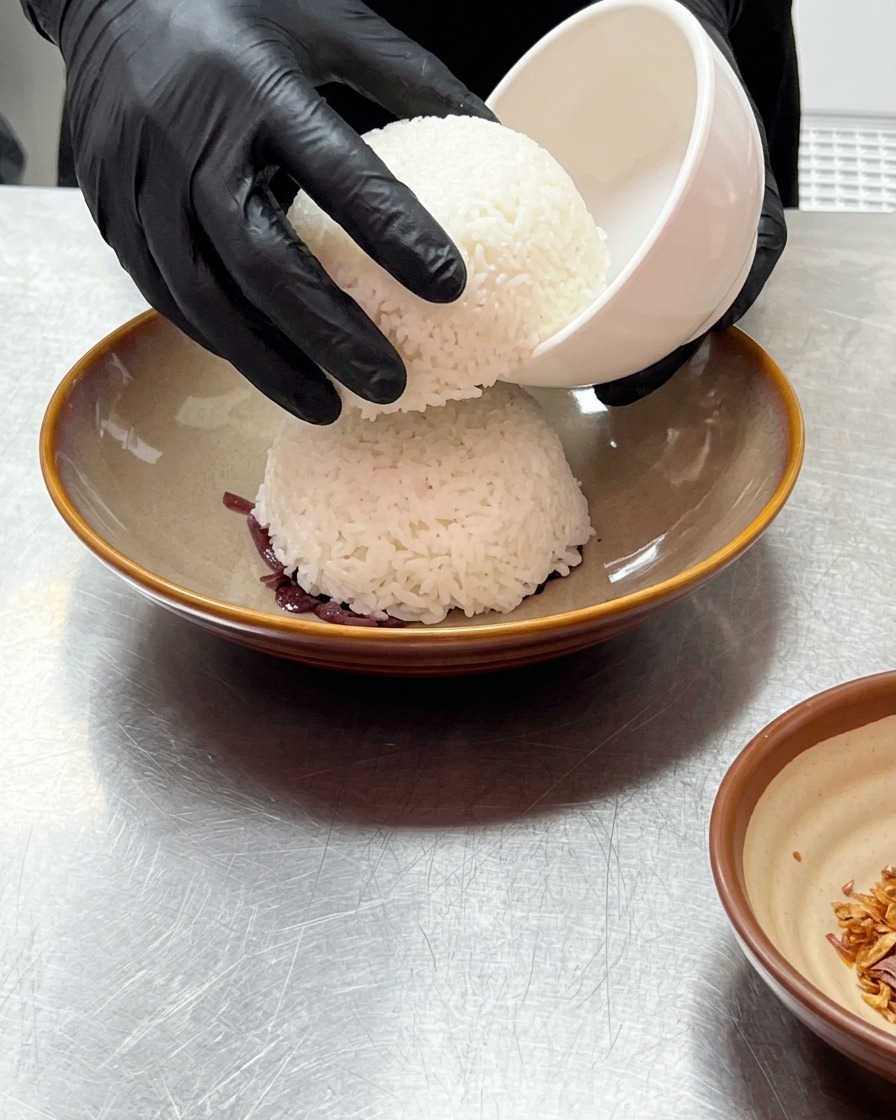 Gloved hands lifting a small white bowl to reveal a dome of steamed white rice resting on top of shredded purple onions.