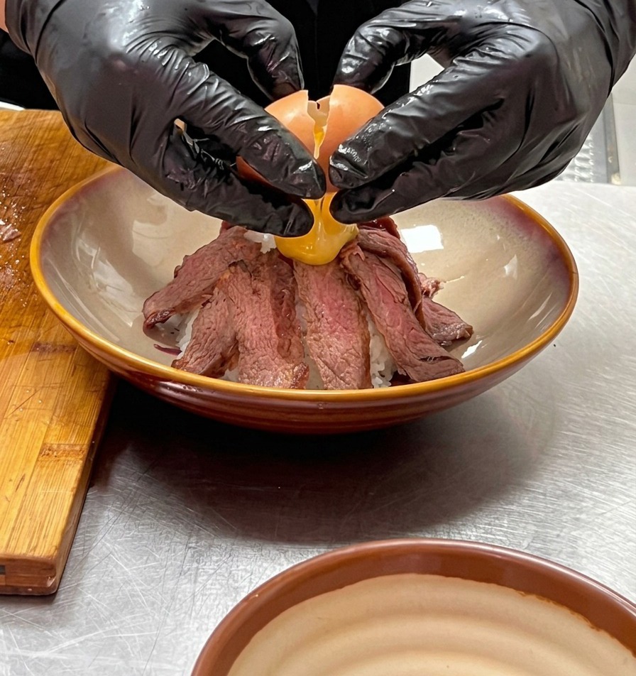 Two gloved hands carefully cracking a partially cooked onsen egg over the peak of a mountain of sliced beef.