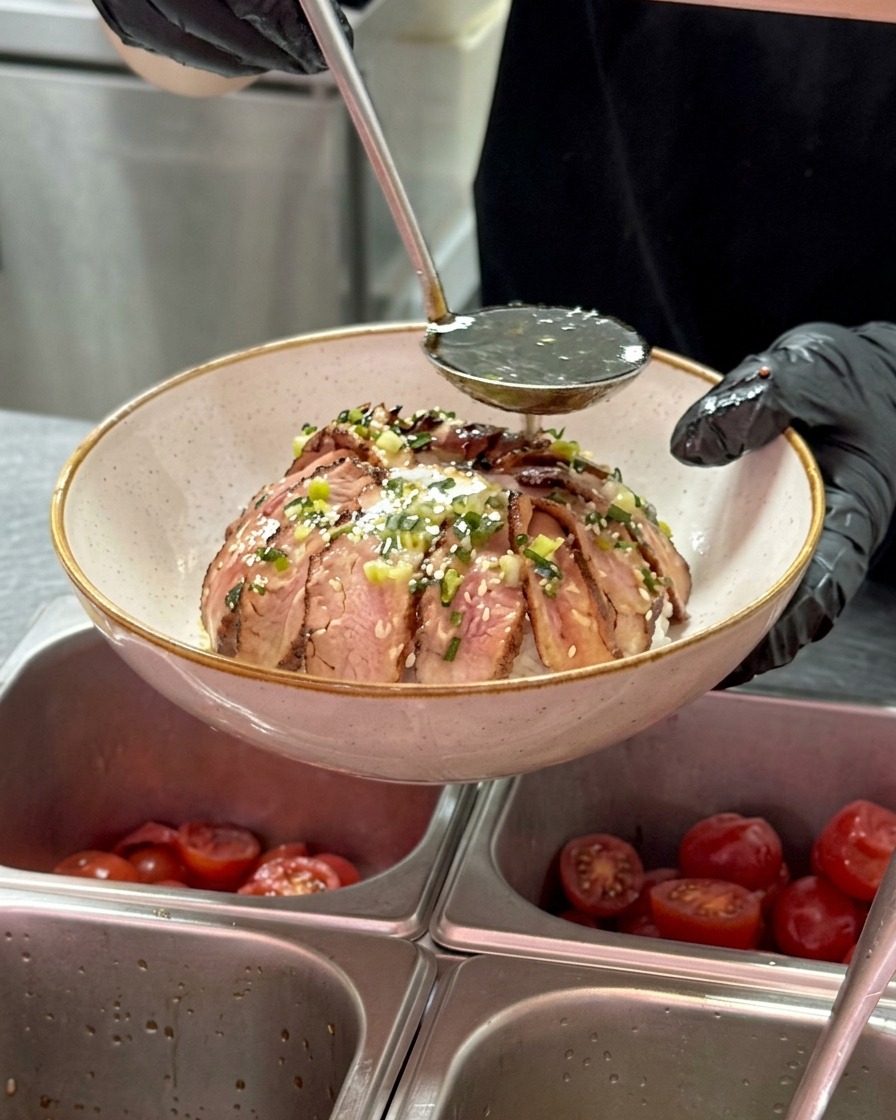 A metal ladle pouring a dark, glossy sauce over a bowl of sliced beef arranged around an egg, with small pieces of scallions visible in the oil.