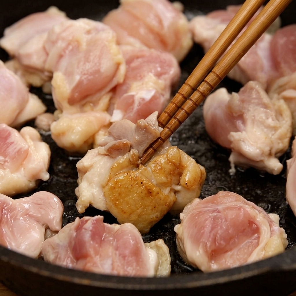 Chicken thigh pieces being stir-fried in a pan with wooden chopsticks until they appear lightly browned.