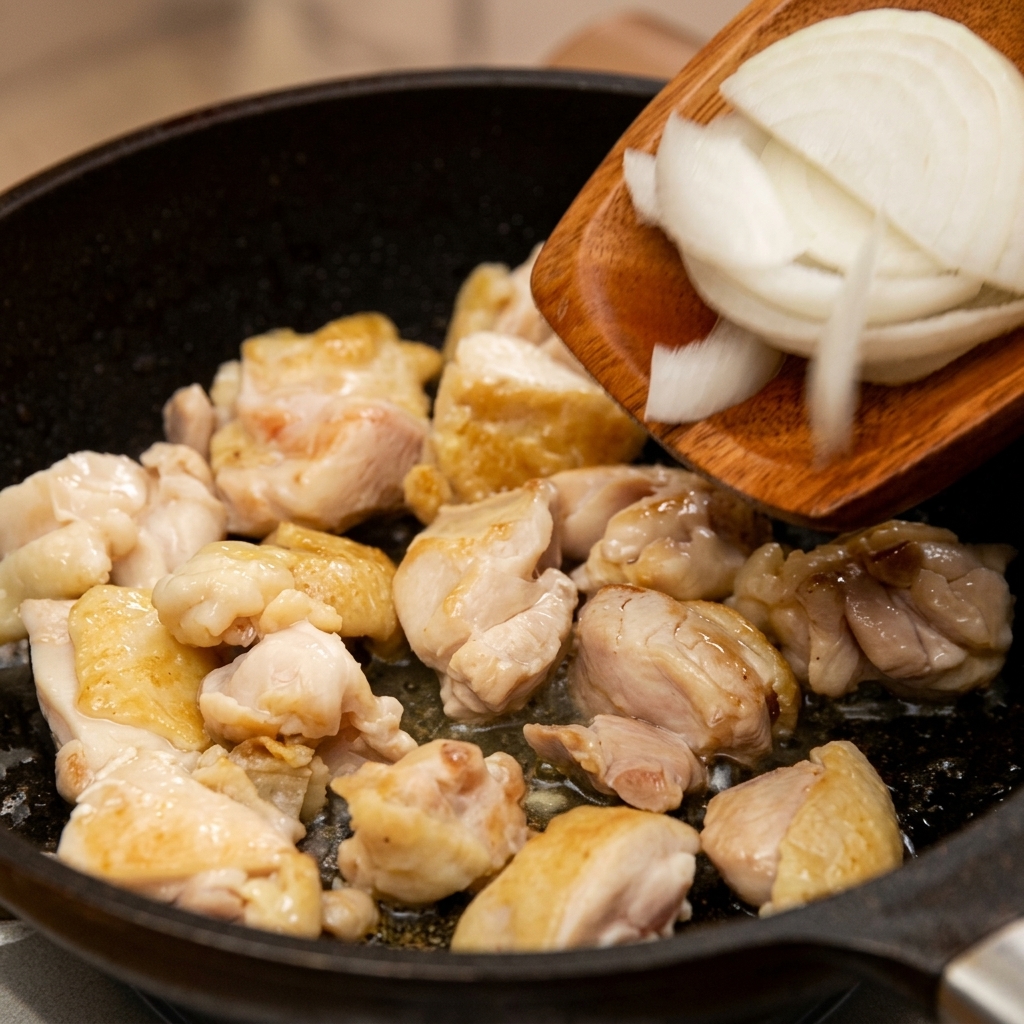 Thinly sliced white onions being transferred from a wooden spatula into a pan with sautéed chicken meat.