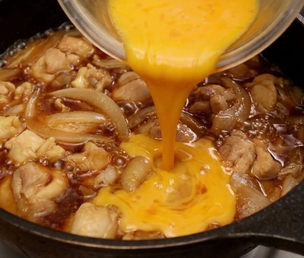 Beaten egg being poured from a glass bowl in a circular stream over chicken and onions in a simmering pan.