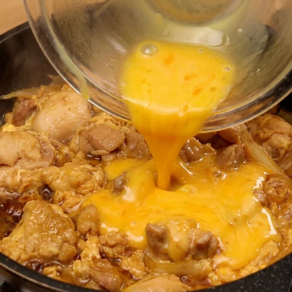 Golden beaten eggs being poured from a glass bowl into a frying pan containing browned chicken and softened onions simmering in a dark sauce.