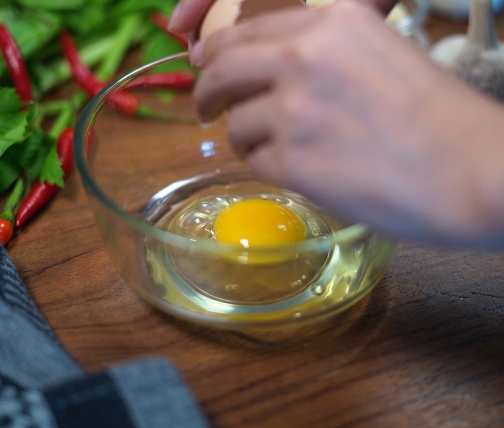 A persons hands carefully cracking a brown eggshell over a clear glass bowl, with a fresh egg yolk and white already visible at the bottom.