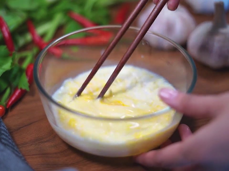 A pair of dark wooden chopsticks stirring a pale yellow mixture of eggs, milk, and cheese inside a glass bowl.