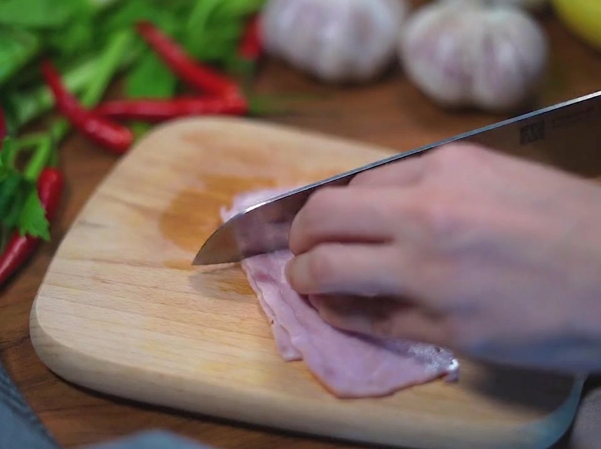 A close-up view of hands using a stainless steel knife to cut strips of pink raw bacon into small squares on a light-colored wooden board.