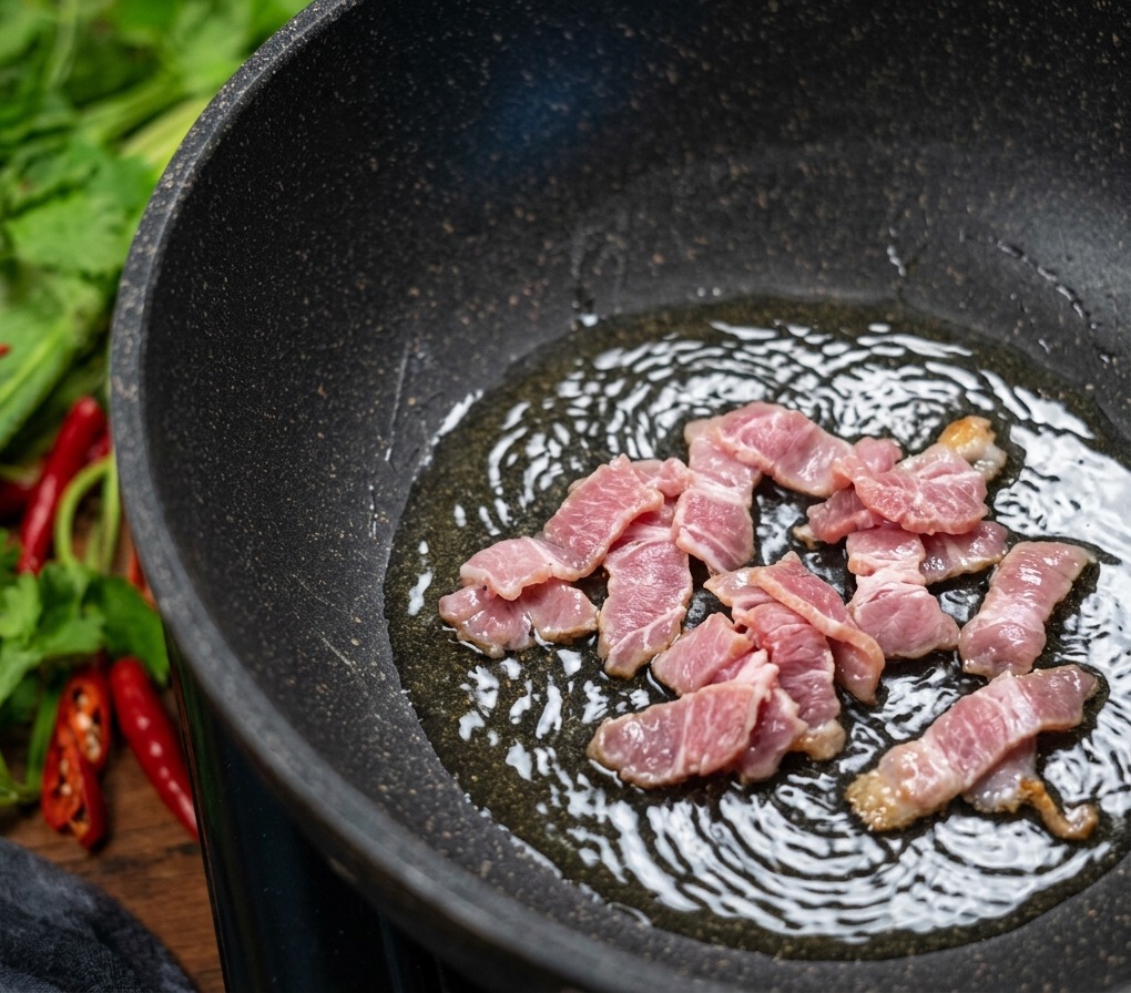 Chopped pieces of bacon sizzling in a dark non-stick pan, with bubbles forming in the rendered fat as the meat browns.