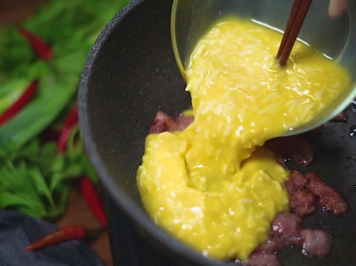 A yellow egg and cheese mixture being poured from a glass bowl into a hot pan containing crispy, browned bacon bits.
