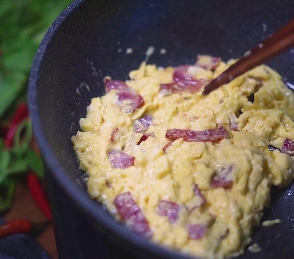 Creamy scrambled eggs mixed with savory bacon pieces being stirred in a dark pan with chopsticks.