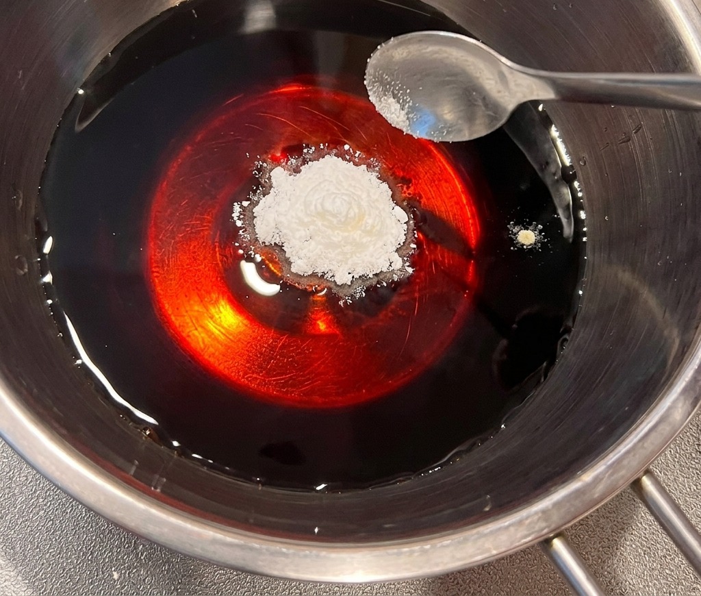 A spoonful of white dashi powder being added to a dark, soy-based liquid in a stainless steel pot.