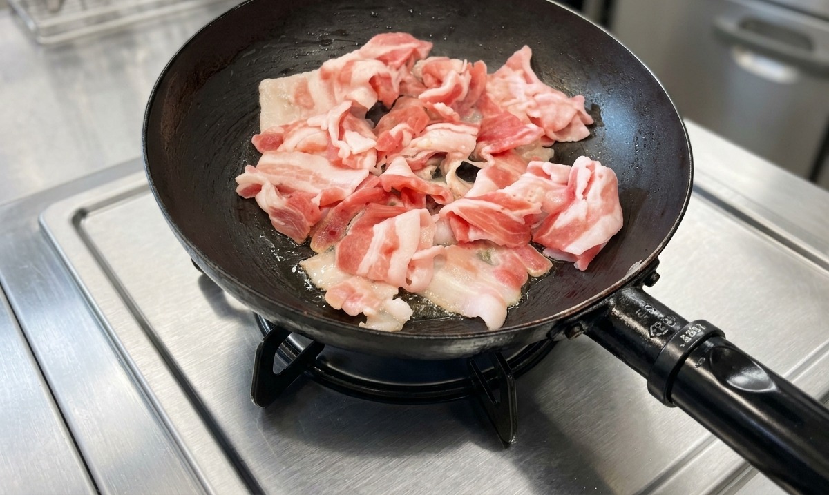 Slices of pork belly being cooked in a black frying pan on a stainless steel stove top.