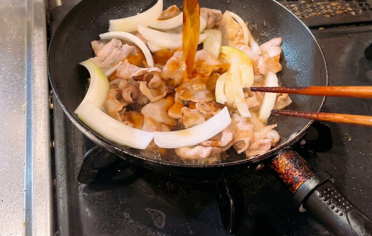 Pouring an amber-colored sauce into a frying pan containing cooked pork belly and raw white onion slices.