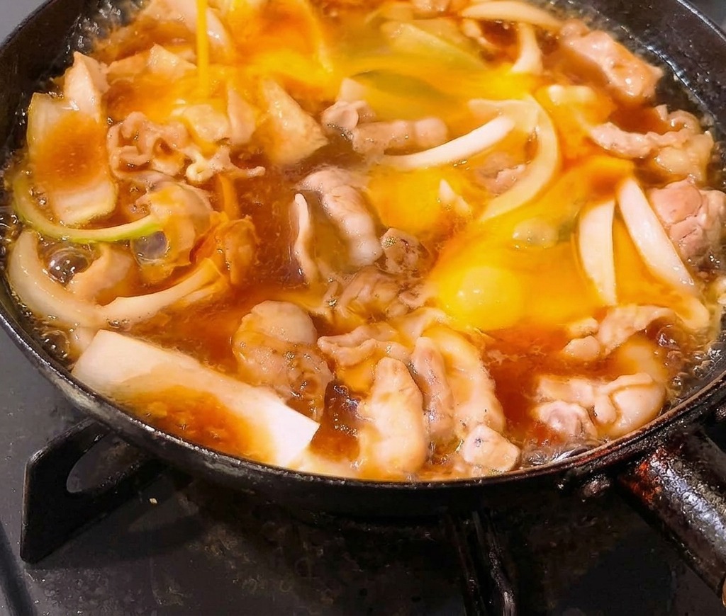Beaten eggs being poured into a frying pan filled with simmering pork belly and onions in a dashi-based sauce.