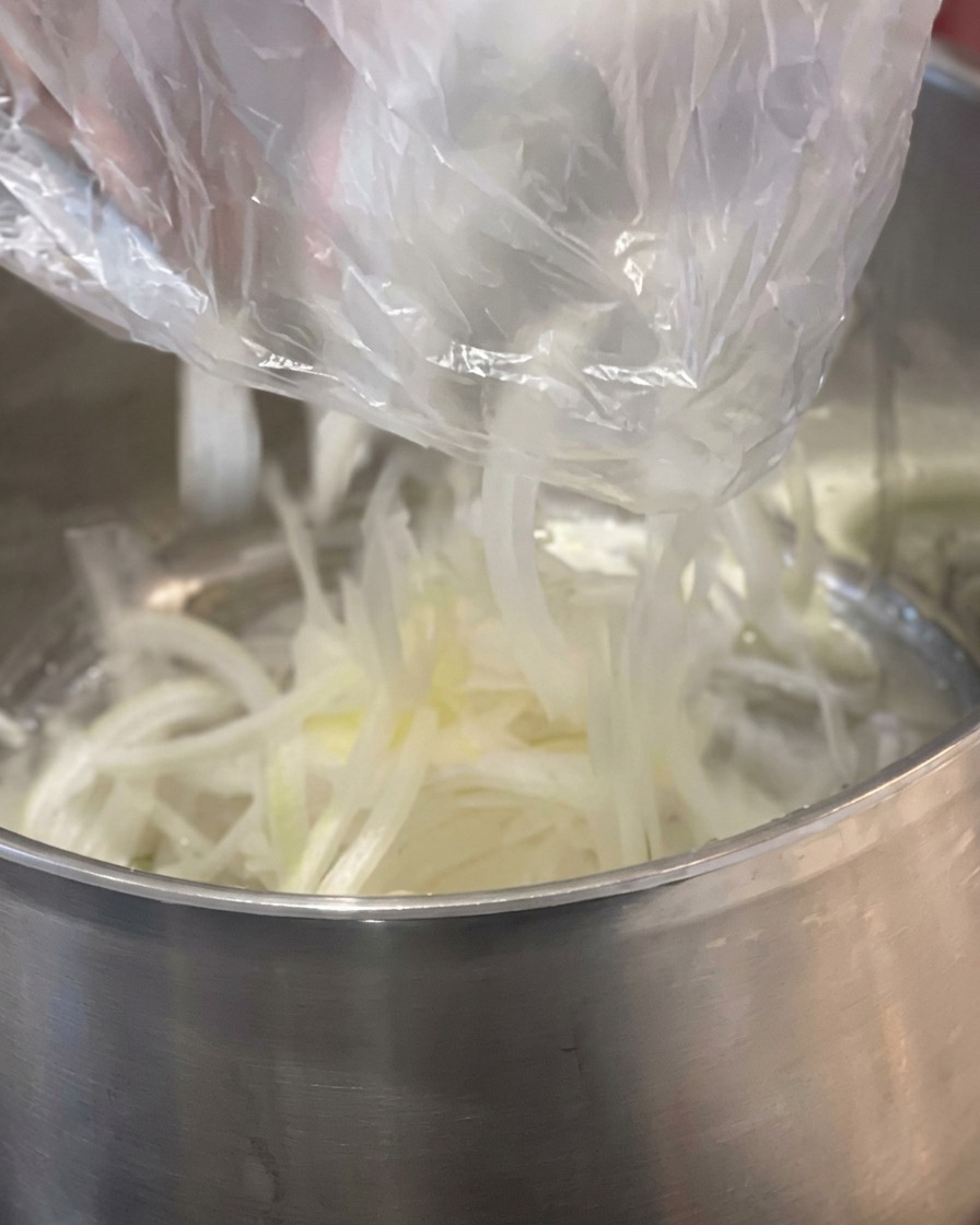 Sliced white onions being dropped from a plastic bag into a stainless steel pot with a block of yellow butter.