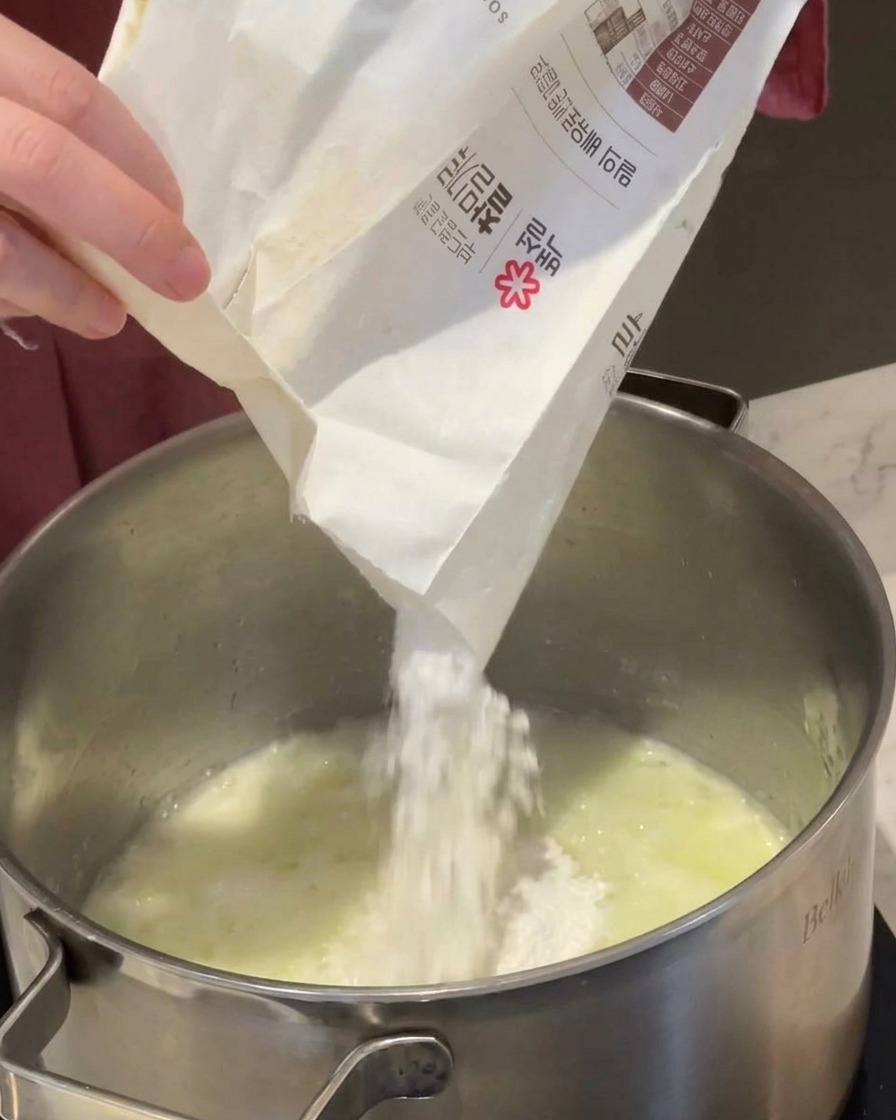 White flour being poured from a paper bag into a pot containing melted butter and onions.