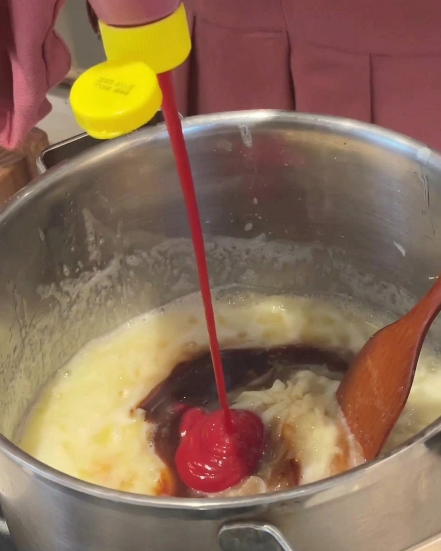Bright red ketchup being squeezed from a yellow-capped bottle into a simmering brown sauce in a metal pot.