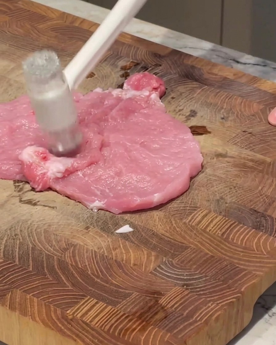 A white meat mallet striking a piece of raw pink pork on a wooden cutting board.