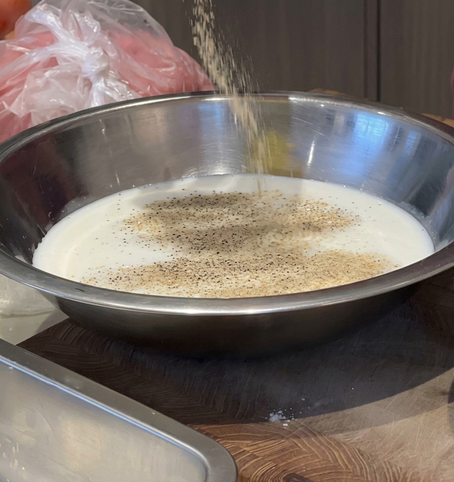 A person pouring seasonings from a container into a silver bowl of milk to create a pork marinade.