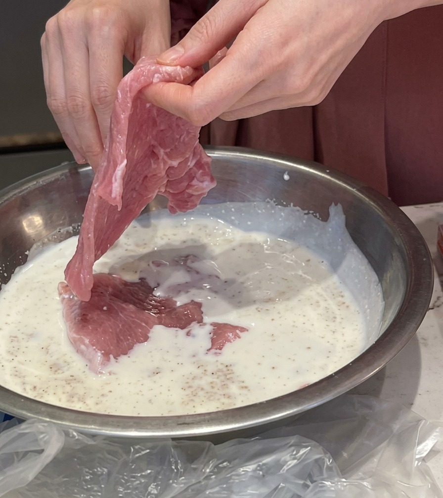 Hands placing a thin slice of raw pink pork into a metal bowl filled with a white milk and garlic marinade.