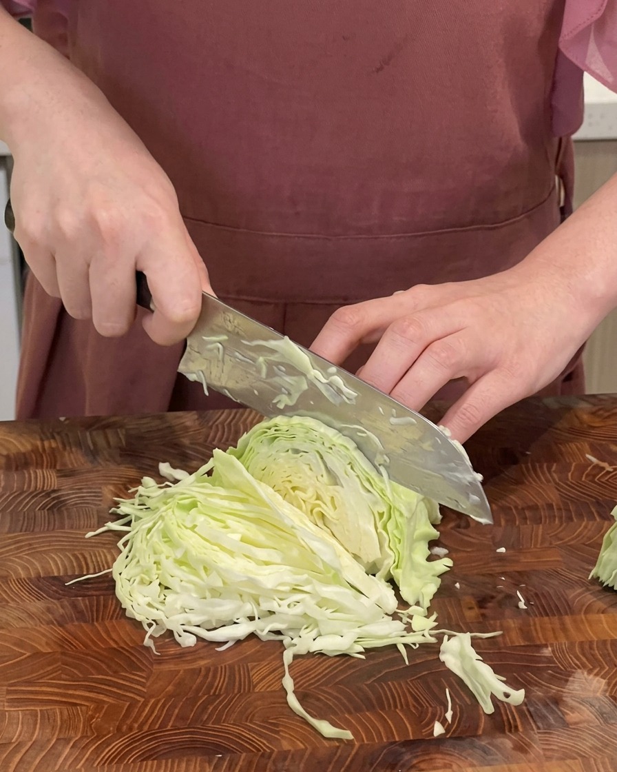A close-up shot of a chef shredding a green cabbage into thin strips on a wooden cutting board with a large knife.