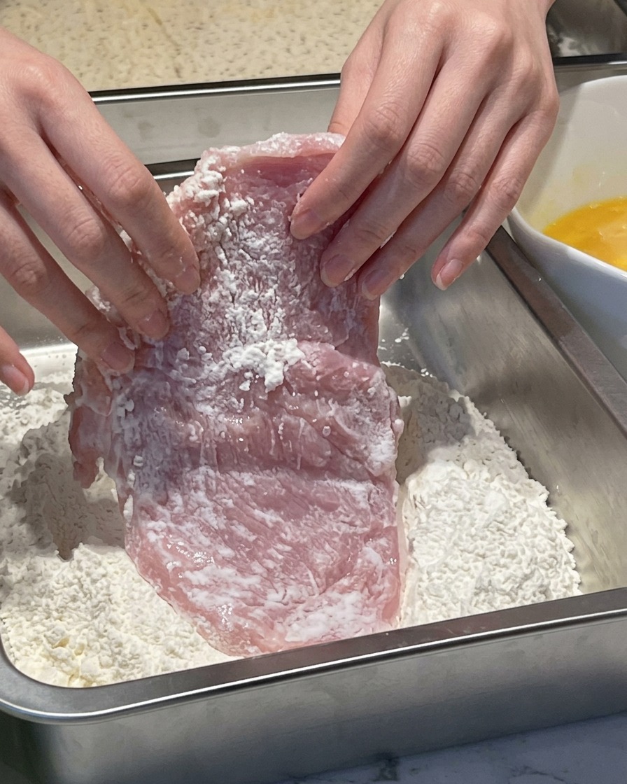 Hands pressing a wet piece of raw pork into a metal container filled with white flour.