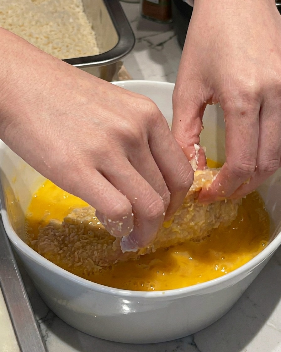 A hand dipping a flour-dusted pork cutlet into a white ceramic bowl containing whisked yellow eggs.