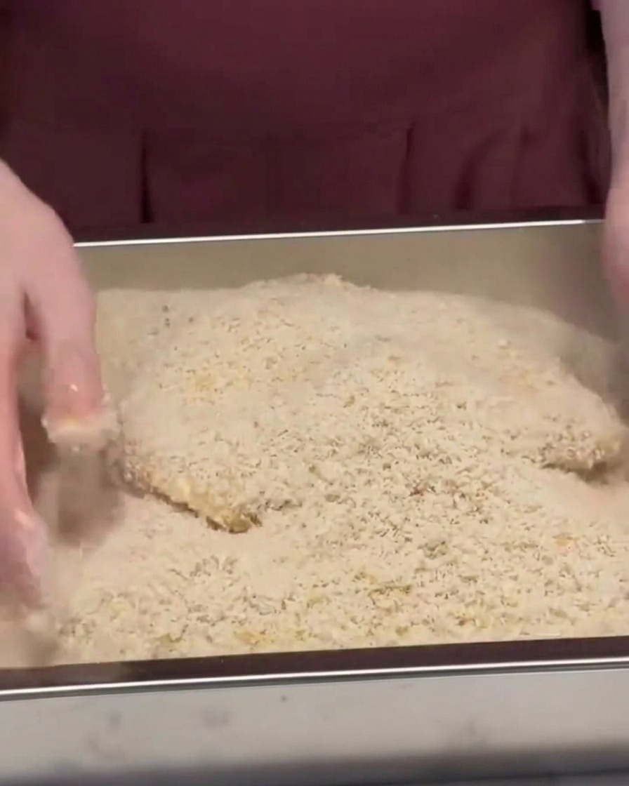 A pork cutlet being buried and pressed into a metal tray filled with panko breadcrumbs.