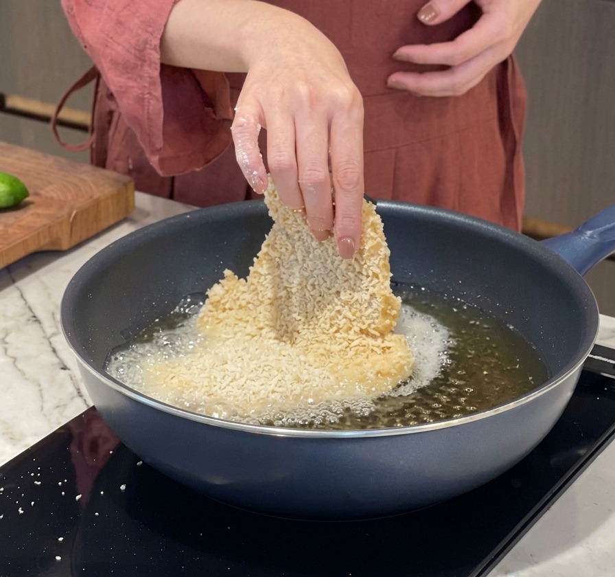 A person hand carefully lowering a large breaded pork cutlet into a hot pan of bubbling oil.